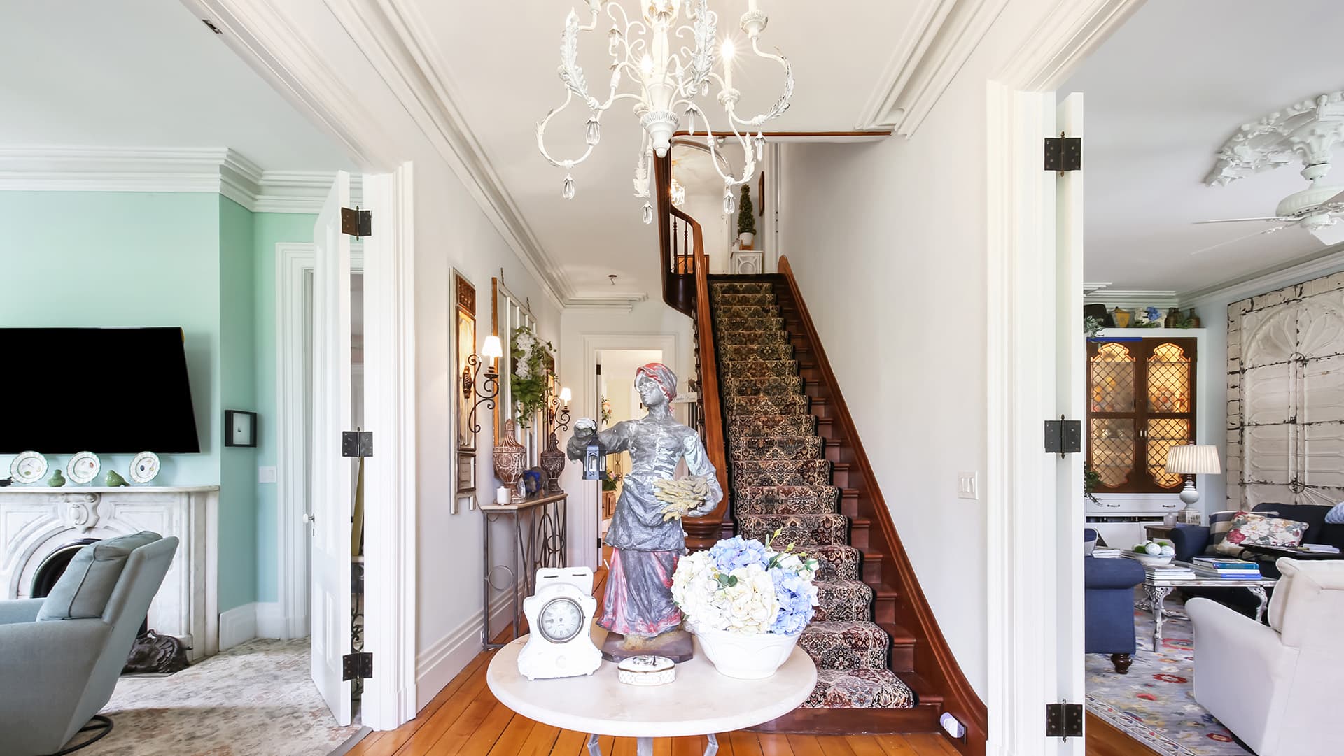 A foyer with a floral centerpiece, a staircase, and a chandelier, opening to a living area.